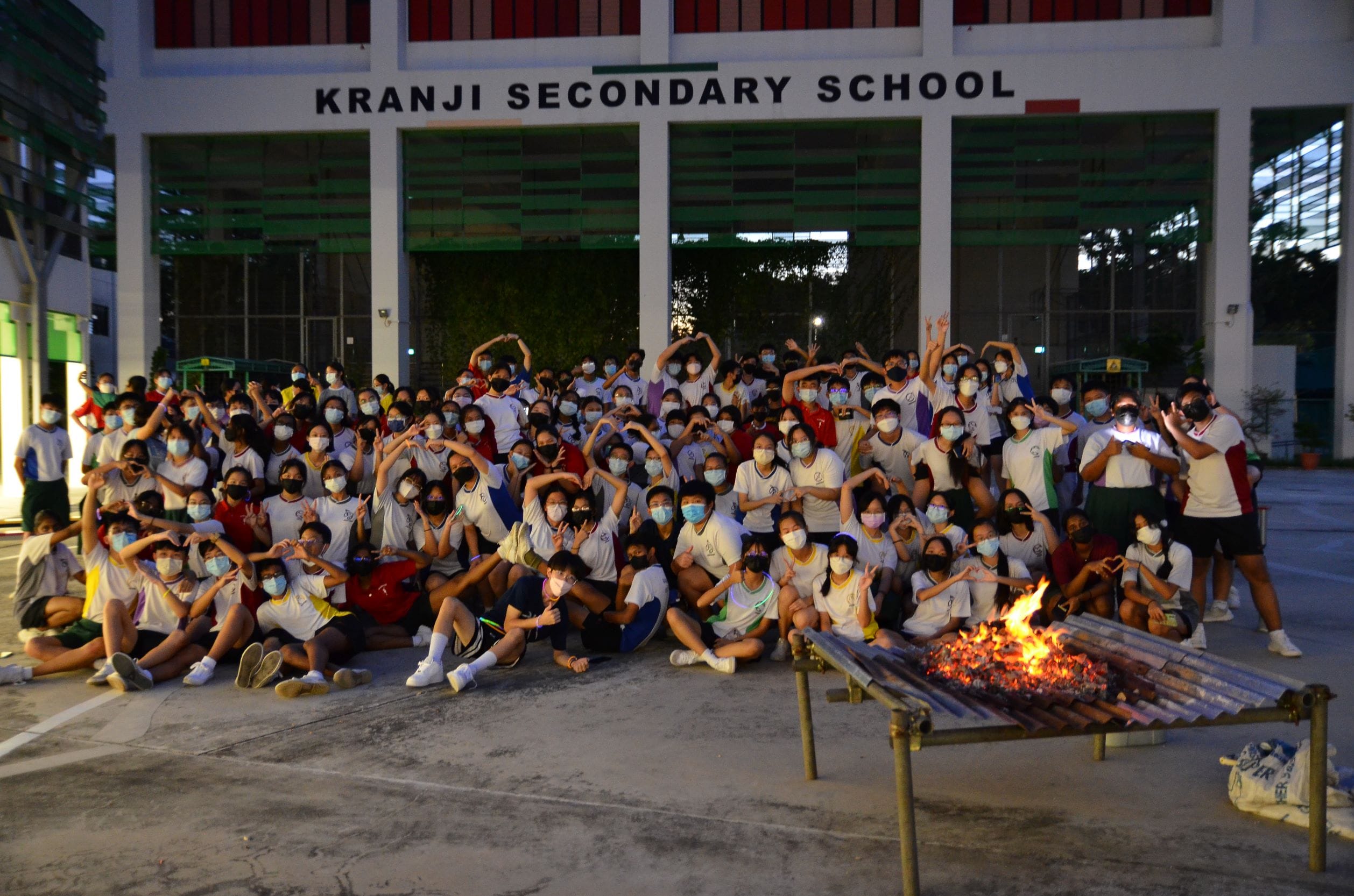 Large masked student group poses in front of Kranji Secondary School building at night with a bonfire in front.
